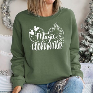 May include: A green sweatshirt with a white graphic design of Mickey Mouse ears with a castle and fireworks. The text on the sweatshirt reads "Magic Coordinator".