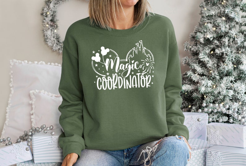 May include: A green sweatshirt with a white graphic design of Mickey Mouse ears with a castle and fireworks. The text on the sweatshirt reads "Magic Coordinator".