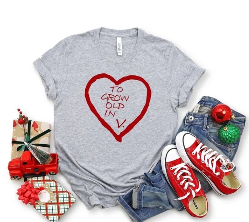 May include: A heather gray t-shirt with a red heart graphic containing the text "TO GROW OLD IN V." is displayed. The shirt is paired with blue jeans and red sneakers. Decorative Christmas ornaments and wrapped gifts are also present.