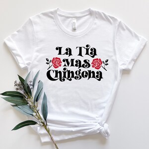 May include: White t-shirt with black text that reads "La Tia Mas Chingona" with red roses on either side of the text.