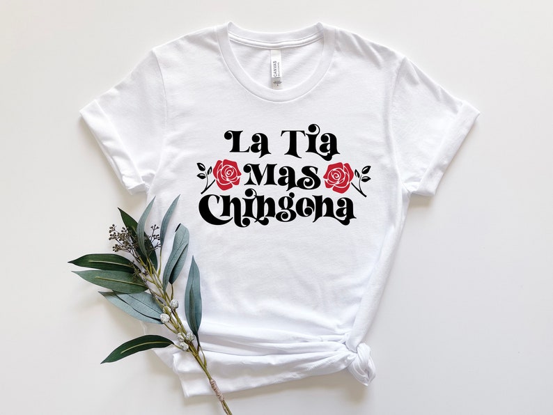 May include: White t-shirt with black text that reads "La Tia Mas Chingona" with red roses on either side of the text.