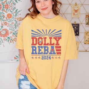 May include: A yellow t-shirt with a vintage-style graphic featuring the text "DOLLY REBA" with a checkmark next to "DOLLY" and a red, white, and blue American flag design. The text "2024" is below the graphic.