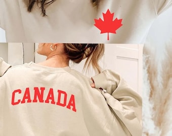 Pode incluir: Uma camisa de moletom bege claro com uma folha de bordo vermelha na frente e a palavra "CANADA" em vermelho nas costas.