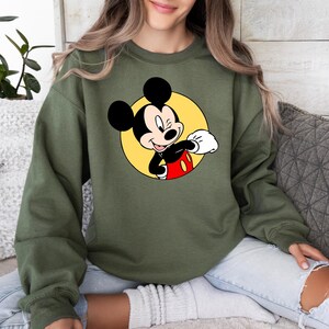 May include: A green sweatshirt with a cartoon image of Mickey Mouse in a yellow circle. Mickey Mouse is wearing red shorts and white gloves and is winking.