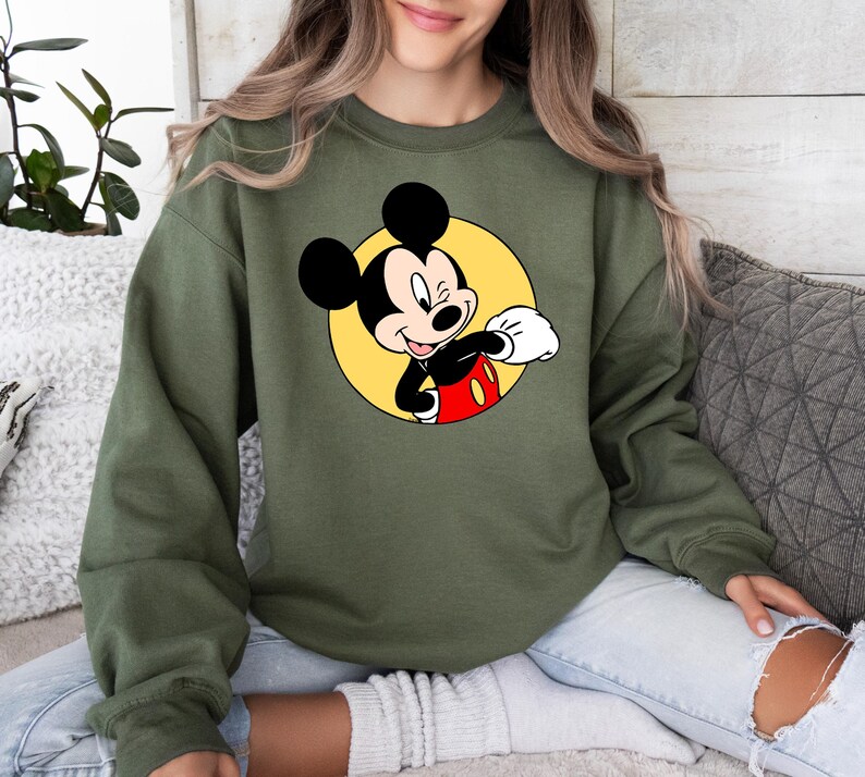 May include: A green sweatshirt with a cartoon image of Mickey Mouse in a yellow circle. Mickey Mouse is wearing red shorts and white gloves and is winking.