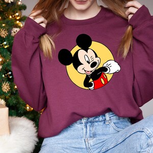 May include: A maroon sweatshirt with a yellow circle and a cartoon image of Mickey Mouse in the center. Mickey Mouse is wearing red shorts and white gloves. He is smiling and waving.