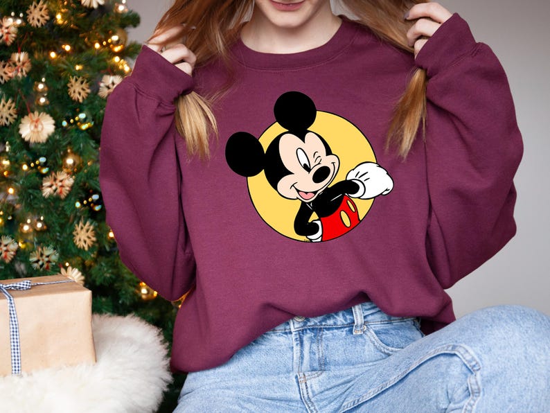 May include: A maroon sweatshirt with a yellow circle and a cartoon image of Mickey Mouse in the center. Mickey Mouse is wearing red shorts and white gloves. He is smiling and waving.
