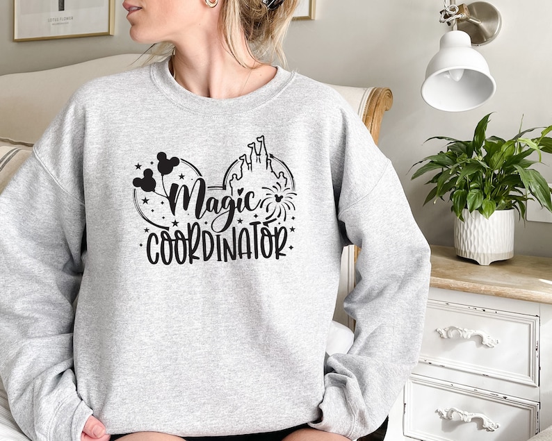 May include: Gray sweatshirt with a black graphic of Mickey Mouse ears, a castle, balloons, and fireworks. The text "Magic Coordinator" is printed below the graphic.