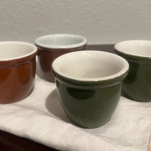 Może przedstawiać: Cztery małe, ceramiczne filiżanki do jajek w odcieniach brązu i zieleni, z białym wnętrzem. Filiżanki ułożone są na białej serwetce, prezentując błyszczące wykończenie i prosty design. Uroczy dodatek do każdej kuchni.