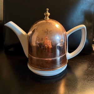 May include: A teapot featuring a white ceramic base, handle, and spout, combined with a polished copper dome-shaped lid. The lid is topped with a small, metallic finial. The reflective copper surface shows a distorted view of the surroundings.