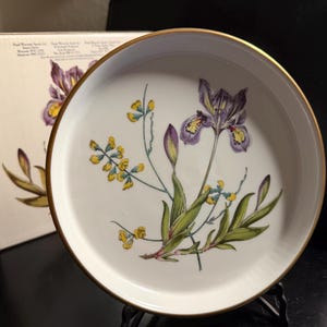 May include: A round white porcelain plate with a gold-toned rim, featuring a botanical illustration of a purple iris and yellow wildflowers with green stems and leaves. The plate is displayed in front of its original white cardboard packaging box.