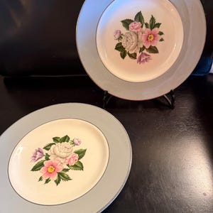 May include: Two round ceramic plates featuring a cream-coloured centre with a floral bouquet of pink, white, and purple flowers with green leaves. The plates have a wide, light blue border and a thin silver-toned rim around the outer edge.