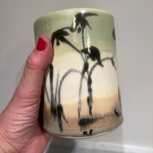 Pode incluir: Uma caneca de cerâmica com um esmalte verde, branco e marrom. A caneca tem um desenho em preto e branco de plantas de bambu no lado. A caneca é assinada na parte inferior com as iniciais "MR".