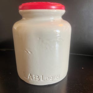 May include: Off-white ceramic jar with a red lid. The jar has a cylindrical shape with a slightly wider base and a narrower neck. The text "LAB Lagny" is embossed on the front of the jar.