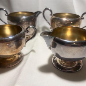 May include: Four antique silver-plated serving pieces, including two handled sugar bowls and two cream pitchers. The interiors have a gold-colored finish, and the exteriors show signs of age. The pieces are on a white surface.