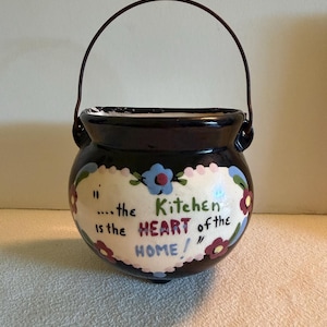 May include: A dark brown ceramic pot with a metal handle, featuring a white heart-shaped centre painted with blue and red flowers. The text reads: "...the Kitchen is the HEART of the HOME!" The pot has a rounded body and a wide opening at the top.