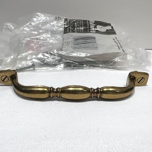May include: A brass-toned cabinet pull with a curved handle and square end pieces. The handle has a series of rounded sections. A screw is visible near the handle. Hardware is in a plastic bag in the background.