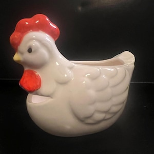 May include: A ceramic hen-shaped planter, painted white with red accents on the comb and wattle. The planter has a hollowed-out area for holding small plants or decorative items. The design is a charming addition to any home decor.