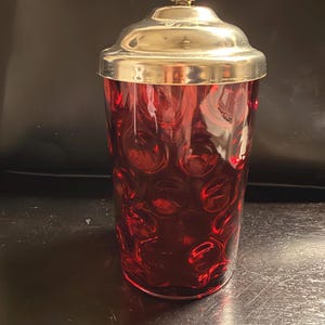 May include: A cylindrical red glass canister featuring a textured bubble pattern on the exterior. The container is topped with a polished silver-toned metal lid that includes a decorative, ornate handle at the very top center.