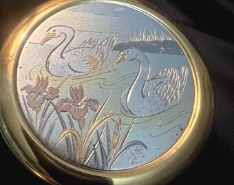 Könnte beinhalten: Ein runder, goldfarbener Dekorteller mit einem silbernen und goldenen Design von zwei Schwänen, die in einem Teich mit Iris schwimmen. Der Teller hat eine strukturierte Oberfläche und einen erhöhten Rand.
