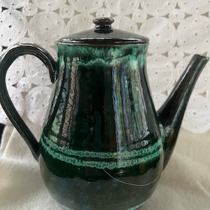 May include: A dark green ceramic teapot with a light green glaze and a brown rim. The teapot has a rounded body and a spout on the right side.