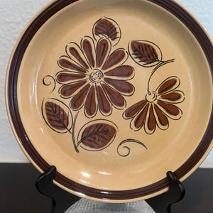May include: A brown and tan ceramic dinner plate with a floral design. The plate has a brown rim and a brown floral pattern on a tan background.