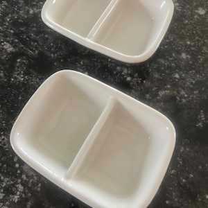 以下が含まれることがあります： 白の長方形の仕切り付きサービング皿が2つ。各皿には、隆起した仕切りで区切られた2つのコンパートメントがあります。皿は光沢のある滑らかな素材でできており、暗く斑点のある背景に置かれています。