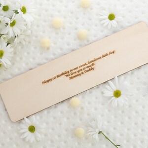 May include: A wooden bookmark with a personalized message: "Happy 1st Birthday to our sweet, handsome little boy! We love you so much! Mommy & Daddy". The bookmark is surrounded by white daisies and small white pom-poms.