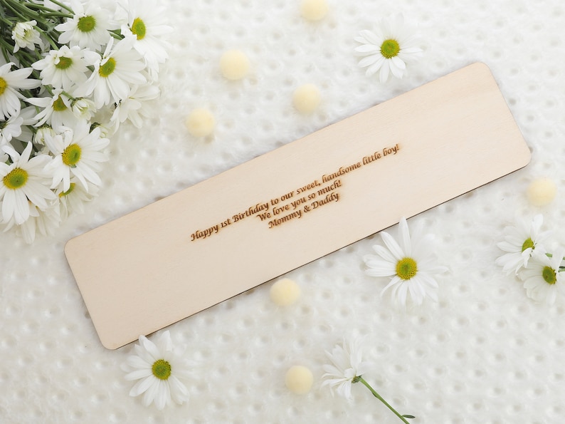May include: A wooden bookmark with a personalized message: "Happy 1st Birthday to our sweet, handsome little boy! We love you so much! Mommy & Daddy". The bookmark is surrounded by white daisies and small white pom-poms.