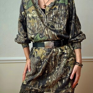 K&ouml;nnte beinhalten: Eine Tunika-Kleid mit Camouflage-Muster, langen &Auml;rmeln und Knopfleiste vorne. Das Kleid ist mit einem schwarzen G&uuml;rtel mit silberner Schnalle in der Taille gerafft. Das Outfit wird mit einer gemusterten Hose kombiniert.
