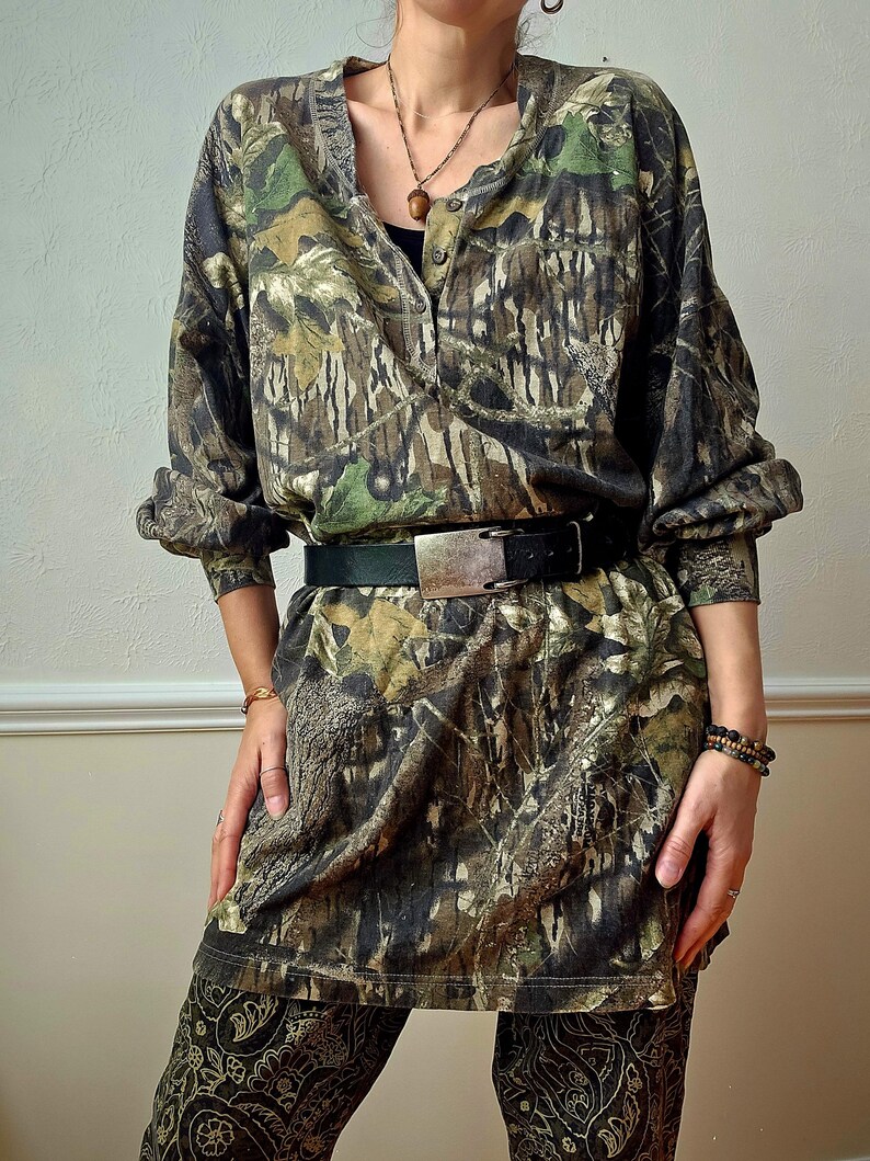K&ouml;nnte beinhalten: Eine Tunika-Kleid mit Camouflage-Muster, langen &Auml;rmeln und Knopfleiste vorne. Das Kleid ist mit einem schwarzen G&uuml;rtel mit silberner Schnalle in der Taille gerafft. Das Outfit wird mit einer gemusterten Hose kombiniert.