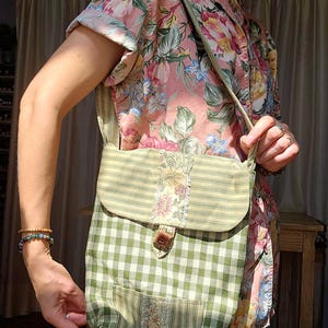 May include: A handmade green and white checkered shoulder bag with a flap closure and a small front pocket. The bag features a striped pattern on the flap and a floral accent. The bag is held by a person wearing a floral shirt.