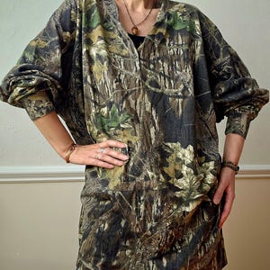 K&ouml;nnte beinhalten: Ein lang&auml;rmeliges Hemd mit Camouflage-Print und V-Ausschnitt. Das Hemd hat ein Muster aus Braun, Gr&uuml;n und Beige, das einem Wald &auml;hnelt. Die &Auml;rmel sind lang und locker, und das Hemd wird mit einer blauen Jeans getragen.