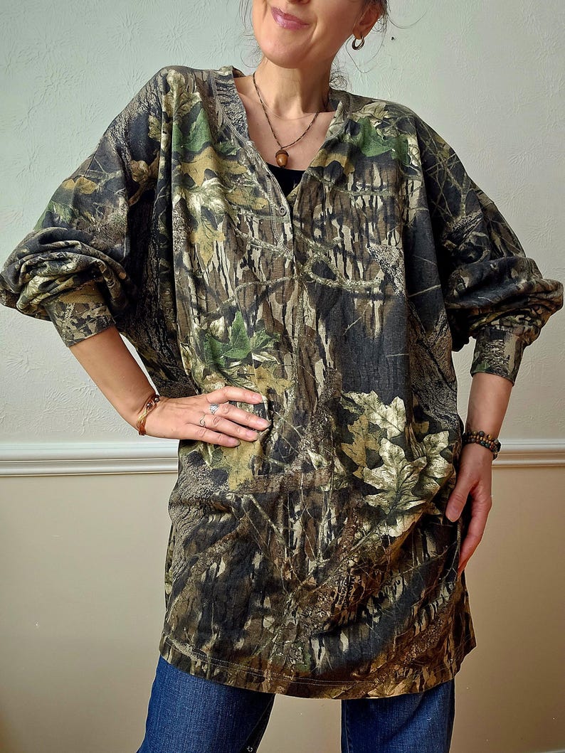 K&ouml;nnte beinhalten: Ein lang&auml;rmeliges Hemd mit Camouflage-Print und V-Ausschnitt. Das Hemd hat ein Muster aus Braun, Gr&uuml;n und Beige, das einem Wald &auml;hnelt. Die &Auml;rmel sind lang und locker, und das Hemd wird mit einer blauen Jeans getragen.