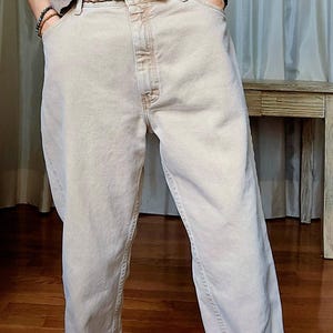 以下が含まれることがあります： ライトベージュのデニムジーンズと茶色の編み込みベルト。ジーンズはクラシックな5ポケットデザインとストレートレッグフィットです。ベルトはシルバー色のバックルが付いています。その人はライトブラウンのTシャツを着ています。