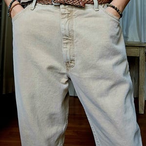 以下が含まれることがあります： 明るいタンデニムのジーンズと、茶色の編み込みレザーベルトのクローズアップ。ジーンズにはフロントポケットとボタン留めが付いています。ベルトにはシルバーのバックルが付いています。その人は明るい茶色のシャツを着ています。