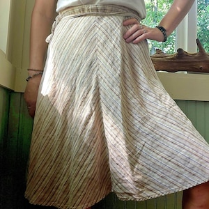 May include: A beige wrap skirt with vertical stripes in shades of brown and blue. The skirt is tied at the waist and falls to the mid-calf. The person is wearing a white t-shirt with a graphic print.