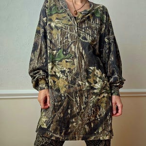 K&ouml;nnte beinhalten: Ein lang&auml;rmliges Hemd mit Camouflage-Print und Knopfleiste. Das Hemd weist eine Mischung aus Gr&uuml;n-, Braun- und Schwarzt&ouml;nen auf, wodurch ein Waldmuster entsteht. Die &Auml;rmel sind lang und locker. Das Hemd wird mit einer gemusterten Hose kombiniert.