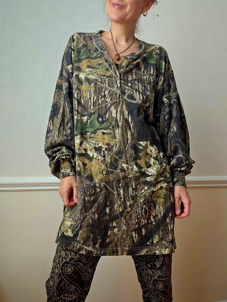 K&ouml;nnte beinhalten: Ein lang&auml;rmliges Hemd mit Camouflage-Print und Knopfleiste. Das Hemd weist eine Mischung aus Gr&uuml;n-, Braun- und Schwarzt&ouml;nen auf, wodurch ein Waldmuster entsteht. Die &Auml;rmel sind lang und locker. Das Hemd wird mit einer gemusterten Hose kombiniert.