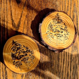 Può includere: Due sottobicchieri rotondi in legno con un disegno di albero inciso. I sottobicchieri sono realizzati con sezioni trasversali di un ramo d'albero, mostrando la venatura naturale del legno e i bordi della corteccia. Il disegno dell'albero è un semplice contorno.
