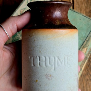 May include: A small, cylindrical ceramic jar with a brown top and the word "THYME" embossed on the front. The jar has a light beige body with an orange gradient near the top. The jar is likely for storing herbs.