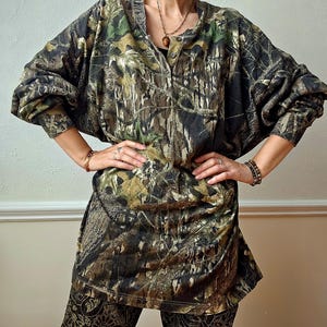 K&ouml;nnte beinhalten: Ein zweiteiliges Set aus Oberteil und Hose mit Tarnmuster. Das Oberteil hat eine Knopfleiste und einen runden Kragen. Die Hose hat ein dunkles Paisley-Design. Die Farbpalette umfasst Gr&uuml;n-, Braun- und Schwarzt&ouml;ne.