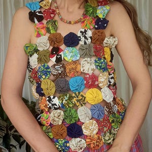 May include: A sleeveless top constructed from numerous small, circular fabric yo-yos in a wide variety of colours and patterns, including floral, polka dot, and solid prints. The garment features an open, grid-like structure connecting the fabric circles.