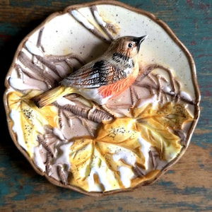 以下が含まれることがあります： 詳細な鳥が枝に止まり、黄色と茶色の葉に囲まれたセラミック装飾プレート。 プレートはクリーム色の背景とテクスチャ加工された縁があります。 鳥は茶色、黒、オレンジ色の羽を持っています。