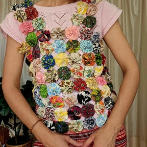 May include: A light pink short-sleeved knit top featuring a dense arrangement of small, circular fabric rosettes in various multicoloured floral, plaid, and geometric patterns covering the front and shoulder areas of the garment.