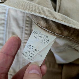 以下が含まれることがあります： ベージュ色のデニムパンツのクローズアップ。タグには「MSR01CE」や「427-00% COTTON」などの文字が表示されています。パンツはアメリカ製です。