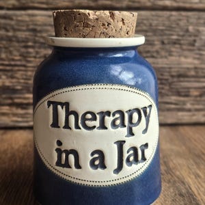 Op de afbeelding: Een blauwe keramische pot met een kurken stopper. De pot heeft een witte ovale vorm met de woorden "Therapy in a Jar" in zwarte letters. De pot staat op een houten oppervlak.