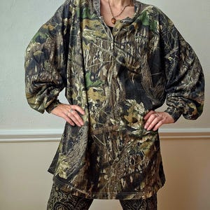 K&ouml;nnte beinhalten: Ein lang&auml;rmeliges Camouflage-Hemd mit Knopfleiste und passender Hose. Das Hemd und die Hose weisen ein Muster aus Braun, Gr&uuml;n und Schwarz auf, das einem Wald &auml;hnelt. Die Hose hat ein Paisley-Design.