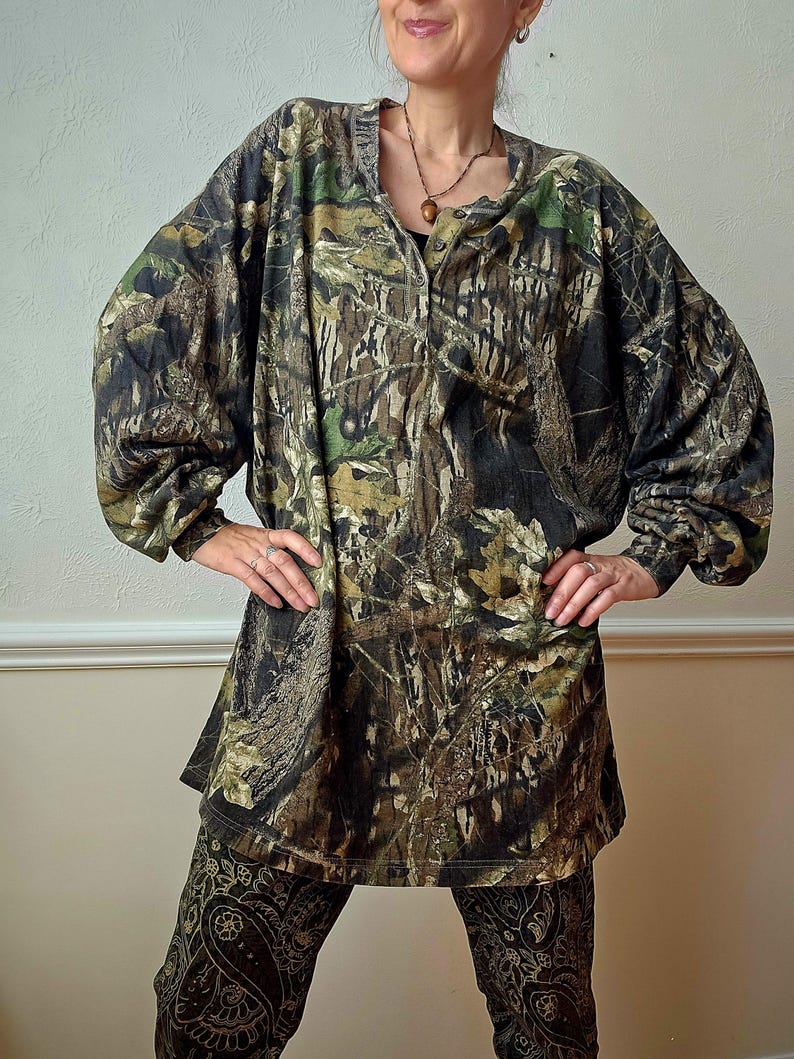 K&ouml;nnte beinhalten: Ein lang&auml;rmeliges Camouflage-Hemd mit Knopfleiste und passender Hose. Das Hemd und die Hose weisen ein Muster aus Braun, Gr&uuml;n und Schwarz auf, das einem Wald &auml;hnelt. Die Hose hat ein Paisley-Design.