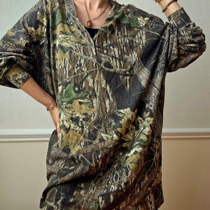 K&ouml;nnte beinhalten: Ein lang&auml;rmeliges Camouflage-Shirt in Braun-, Gr&uuml;n- und Schwarzt&ouml;nen. Das Shirt hat einen V-Ausschnitt und eine lockere Passform. Der Stoff weist ein detailliertes Muster aus Bl&auml;ttern und &Auml;sten auf, das einen Waldeffekt erzeugt. Das Shirt wird mit einer blauen Jeans kombiniert.